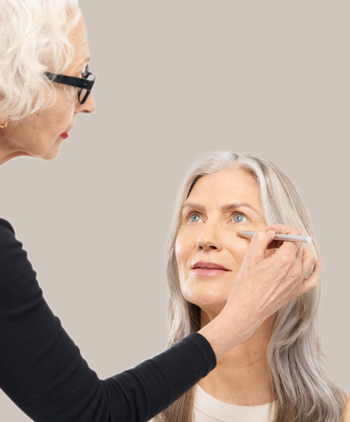 A woman with silver hair has concealer applied under her eye using the Skin2Skin Concealer Brush held by another woman wearing glasses, showcasing a gentle makeup application.