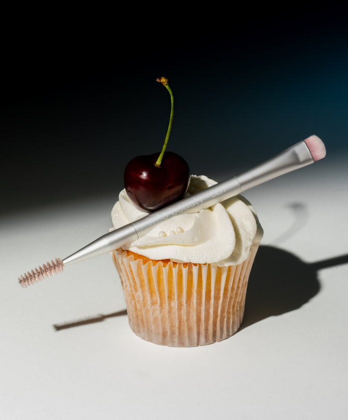 Back2Brow Brush with dual ends featuring a spoolie and angled brush resting on a cupcake topped with white frosting and a cherry, highlighting clean beauty and sustainable packaging.