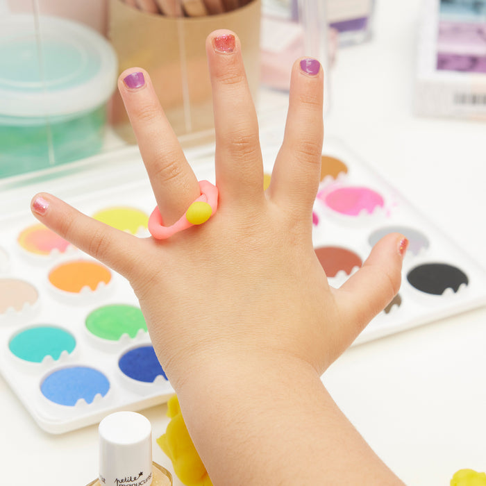 A child's hand with colorful painted nails using Manucurist's Betty Scarlett Stella Trio is shown above a palette of watercolor paints, art supplies, and a pink and yellow ring in a bright, creative workspace.