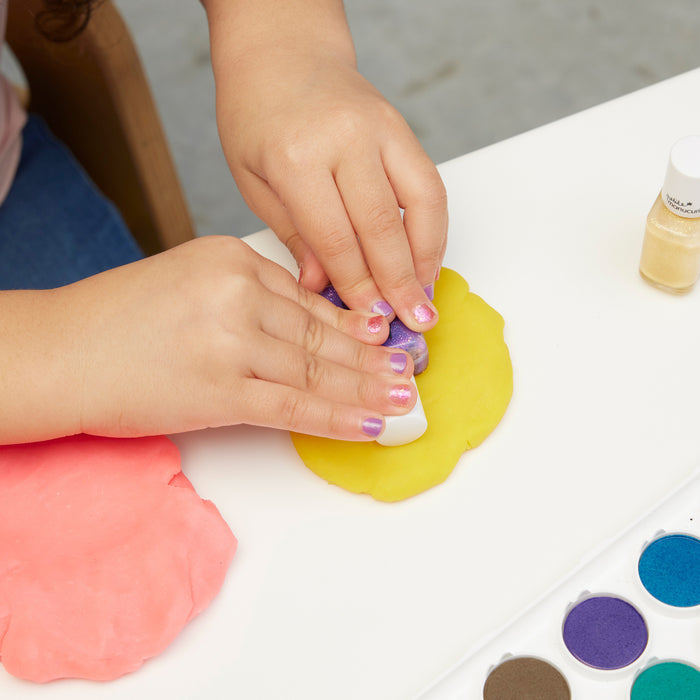 A child with colorful nail polish from the Manucurist Betty Scarlett Stella Trio presses a stamp into yellow playdough on a white table, while pink playdough and watercolor paints are scattered nearby.