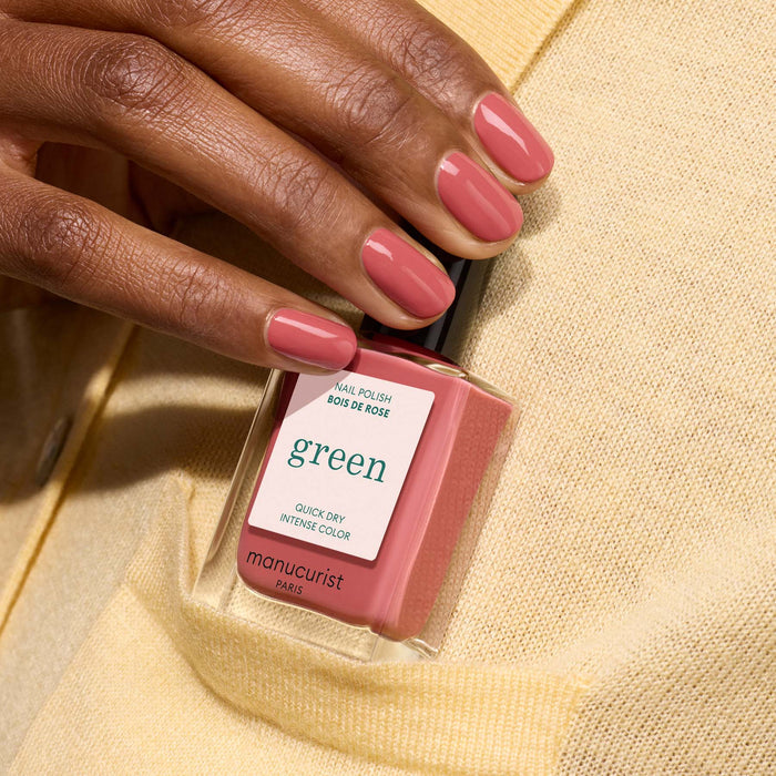 A hand with short, oval nails painted in Manucurist's Bois de Rose holds a matching bottle of polish from the Green™ range against a yellow cardigan.