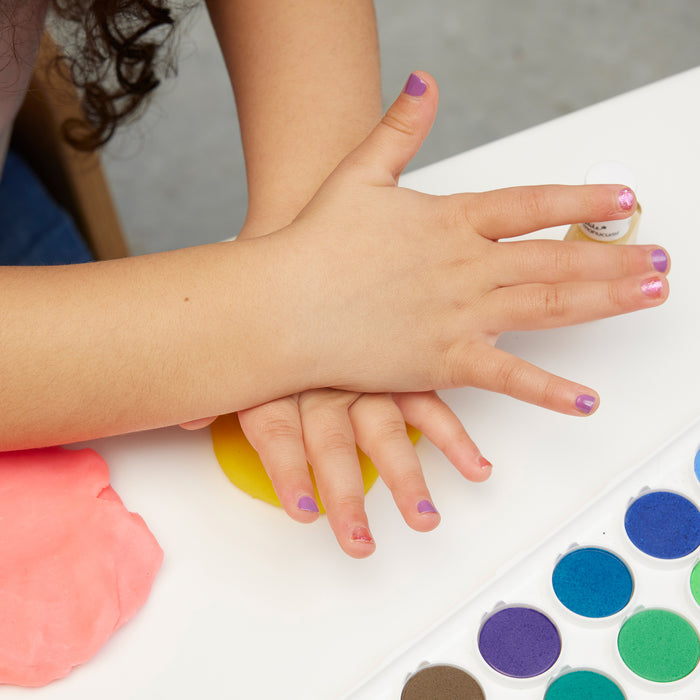 A child with Manucurist's Bonnie Stella Janis Trio non-toxic nail polish presses hands on yellow and pink modeling clay beside a watercolor palette on a white table.