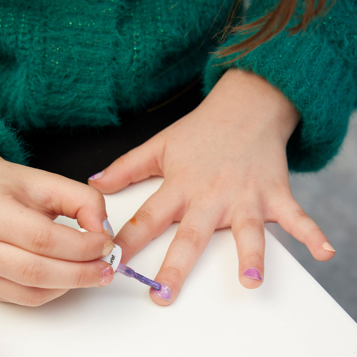 A child in a green sweater sits at a white table, painting their nails with the colorful, non-toxic Bonnie Stella Janis Trio by Manucurist.