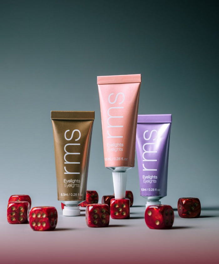 Three tubes of Eyelights Cream Eyeshadow in bronze, peach, and lavender shades arranged among red six-sided dice with gold dots on a neutral background.