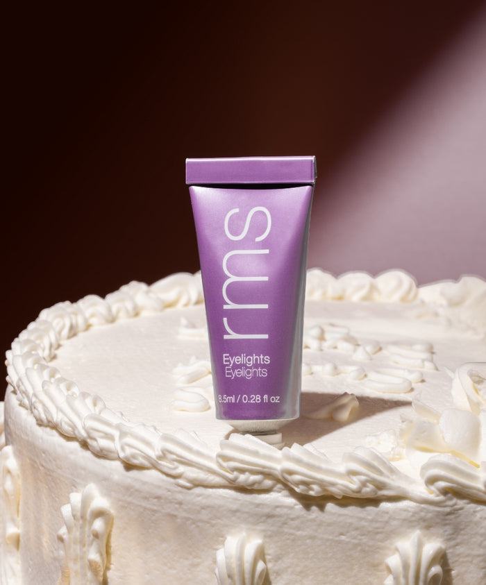 A tube of Eyelights Cream Eyeshadow in a soft purple shade standing upright on a white frosted cake with decorative icing, highlighting clean beauty and natural ingredients.