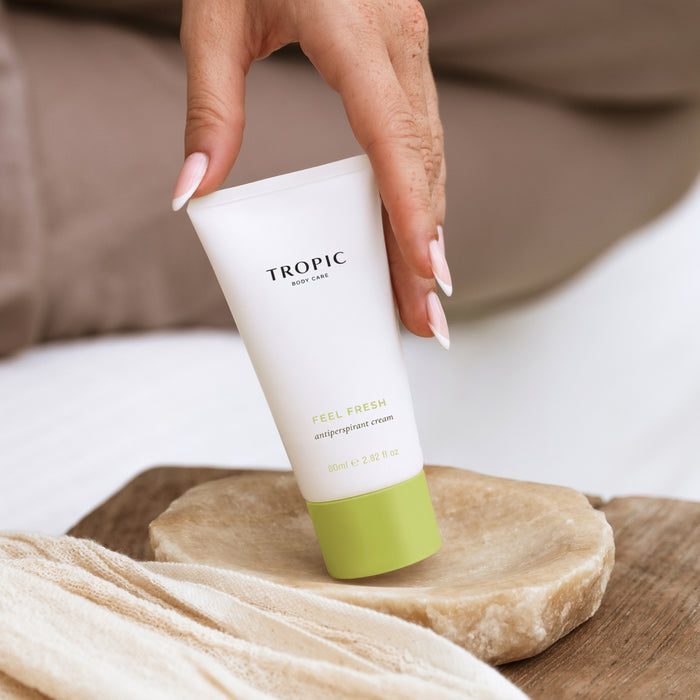 A hand with pale pink manicured nails holds a white tube of Tropic Feel Fresh Antiperspirant Cream, which has a light green cap, resting on a stone dish over a wooden surface.