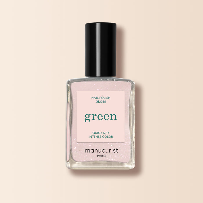 A bottle of Manucurist Gloss nail polish with a black cap and pink label reading "green, quick dry intense color." The iridescent light pink polish inside features subtle shimmer, shown against a beige background.