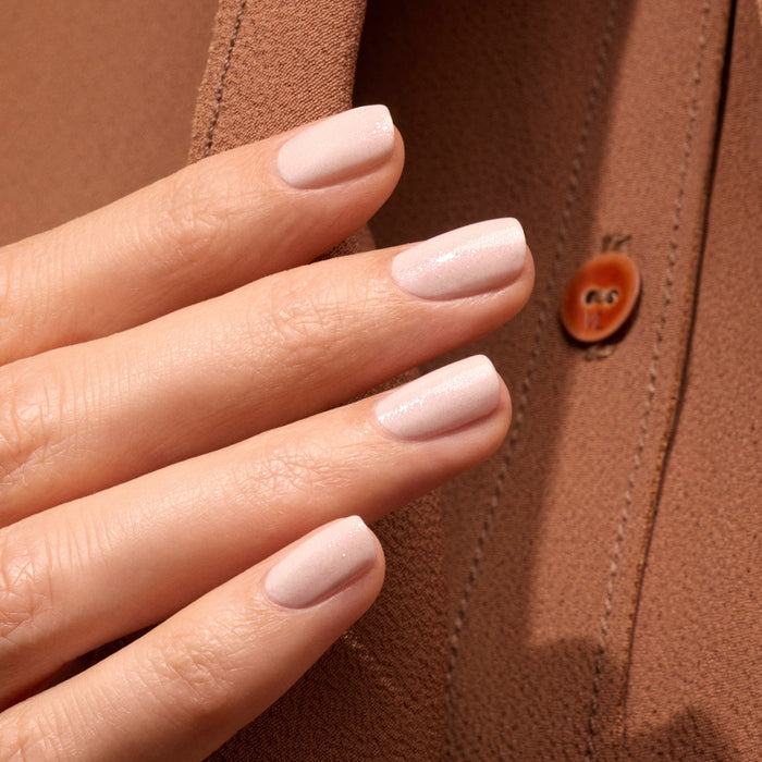 A close-up of a hand with neatly manicured nails painted in Manucurist Gloss, an iridescent pink polish, held against brown fabric with a visible button.