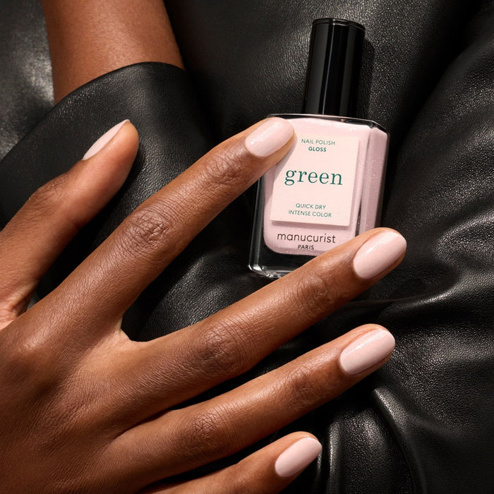 A hand with neatly manicured iridescent pink nails holds a bottle of Manucurist Gloss nail polish against a black leather background.