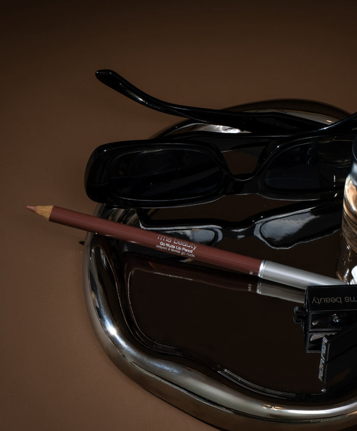Go Nude Lip Pencil displayed on a reflective surface next to black sunglasses and a pencil sharpener, highlighting clean beauty and sustainable packaging.