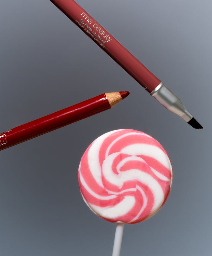 Go Nude Lip Pencil with a pinkish-nude tip and a lip brush next to a pink and white swirled lollipop on a gray background.