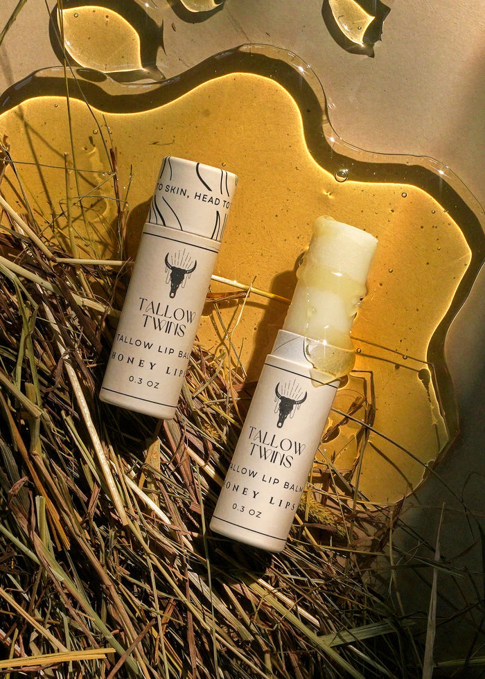 Two Honey Lips tallow lip balm sticks resting on straw beside a pool of honey.