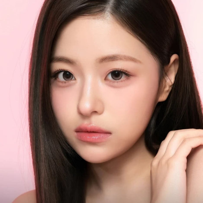 A young woman with long, straight dark hair and fair skin gazes at the camera with a neutral expression against a soft pink background. She wears subtle makeup and is seen wearing i-Sha One Pick Only One 1-Day Cherry Choco contact lenses by i-Sha, which give her eyes a natural dolly effect as she holds her hand near her face.