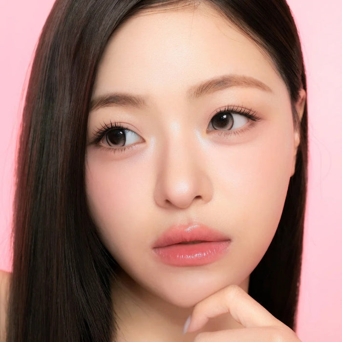 A young woman with smooth, fair skin and long, straight dark hair poses against a pink background, wearing i-Sha One Pick Only One Cherry Choco contact lenses for subtle eye enlargement. Her makeup is natural with glossy lips.