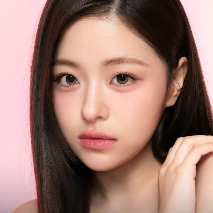 A young woman with long, straight dark hair and clear skin gazes at the camera, her glossy lips and flushed cheeks enhanced by i-Sha One Pick Only One Cherry Choco contacts for a natural eye-enlarging effect against a pink background.