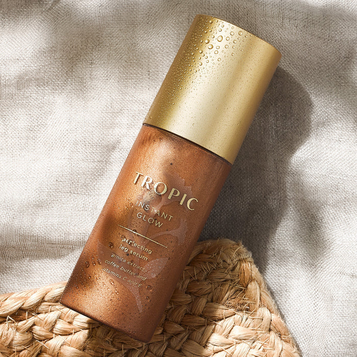 The image features a bottle of "Instant Glow" Perfecting Leg Serum resting on a woven mat, with a soft linen fabric in the background. The bottle has a sleek bronze finish with a golden cap, adorned with water droplets, giving it a fresh and dewy appearance. The product is designed to enhance the skin’s radiance and smoothness, offering a luxurious and sun-kissed glow. The natural and cozy setting evokes warmth and relaxation, aligning with the product's summer-ready theme.