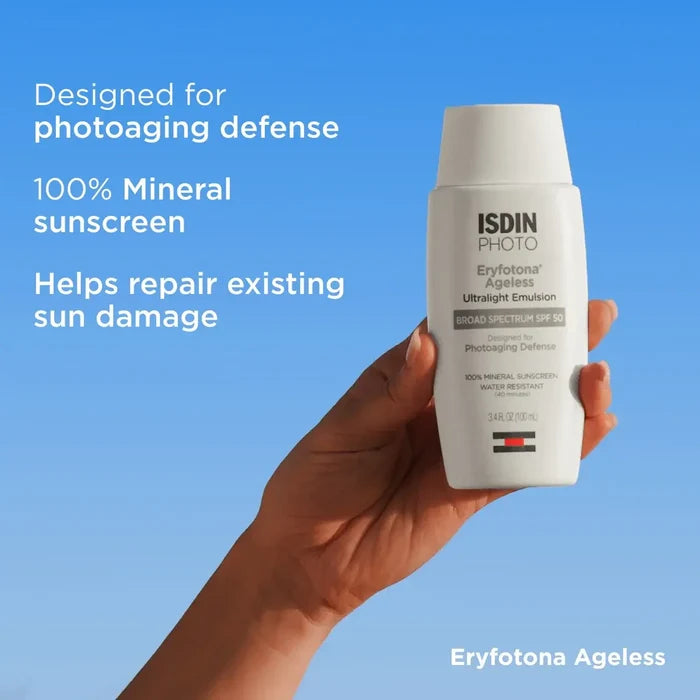 Hand holding ISDIN Eryfotona Ageless mineral sunscreen bottle with SPF 50 on blue background