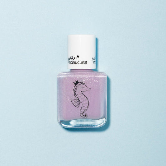 A bottle of Manucurist Janis the Seahorse kids' nail polish in light purple with a white cap, featuring a black crowned seahorse illustration. Made with naturally sourced ingredients and shown on a light blue background.