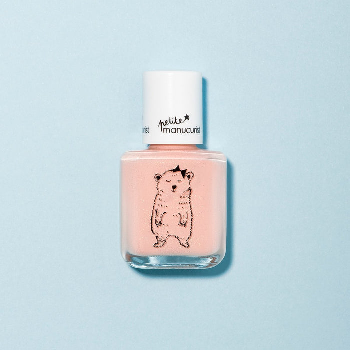 A bottle of Manucurist Joy the Bear Cub light pink kids' nail polish with a white cap and a cute standing bear illustration. Set on a solid light blue background. Made with naturally sourced ingredients for gentle, safe fun.