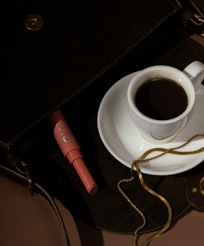 Legendary Serum Lipstick in warm nude shade resting inside an open dark leather bag next to a white cup of black coffee on a saucer and a gold chain necklace, evoking a sophisticated clean beauty vibe.