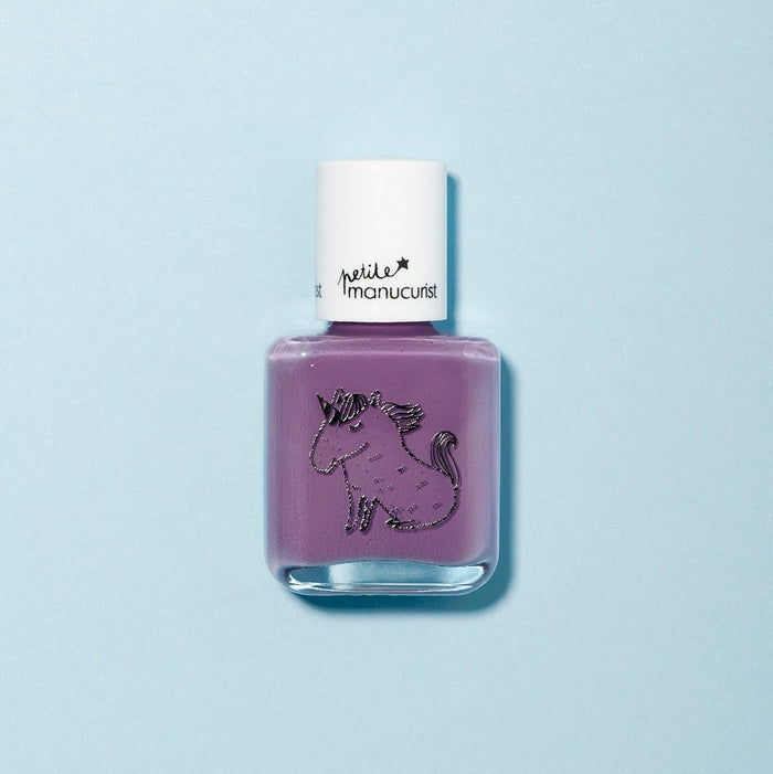 A bottle of Manucurist’s Lily the Unicorn kids nail polish in vibrant purple rests on a light blue background, featuring a unicorn design and white cap, showcasing its fun, naturally sourced formula.