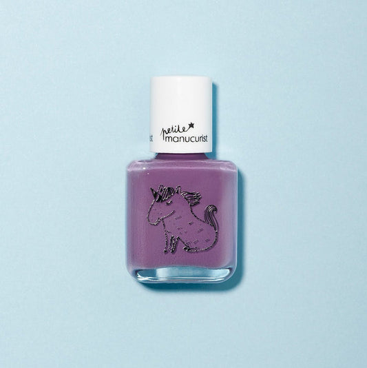 A bottle of Manucurist’s Lily the Unicorn kids nail polish in vibrant purple rests on a light blue background, featuring a unicorn design and white cap, showcasing its fun, naturally sourced formula.