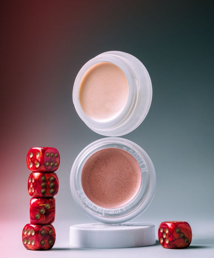 Two open containers of Lip2Cheek cream makeup shown with stacked and scattered red dice, highlighting the smooth texture and natural shades with clean beauty appeal.