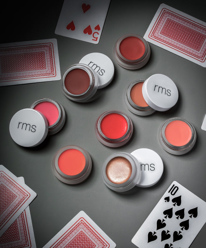 Various shades of Lip2Cheek cream blush in small jars with white lids, displayed alongside playing cards on a gray surface.