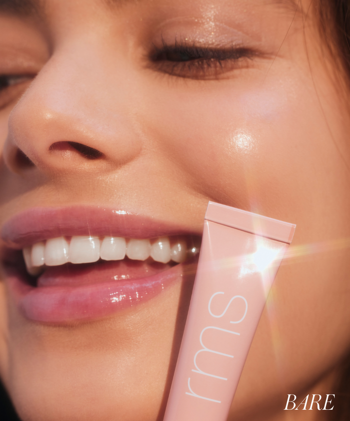 Close-up of a smiling woman with glowing skin holding a pink tube of Liplights Cream Lip Gloss near her cheek in natural light, highlighting smooth and radiant makeup.