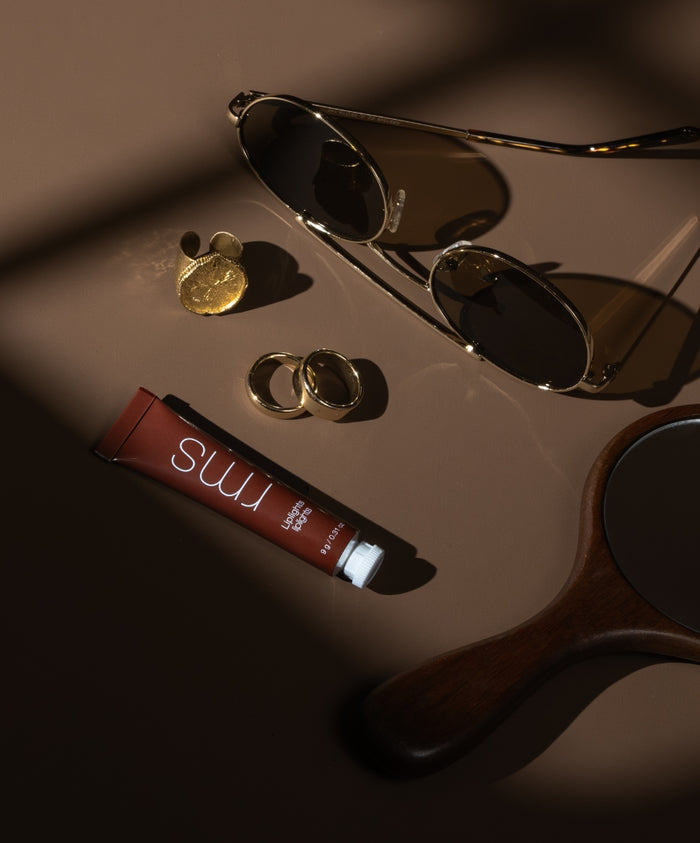 Liplights Cream Lip Gloss tube lying next to gold rings, coin, round sunglasses, and a wooden hand mirror on a soft brown surface in warm natural light.