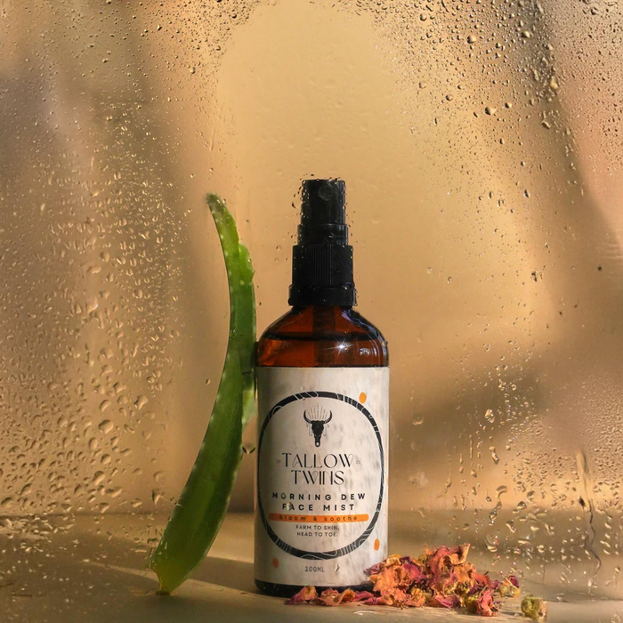 A bottle of Morning Dew Face Mist against a misted window, with an aloe leaf and dried rose petals.
