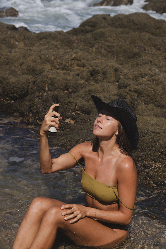 A woman in a black cowboy hat sitting in a rocky tide pool, spraying Tallow Twins mist onto her face.
