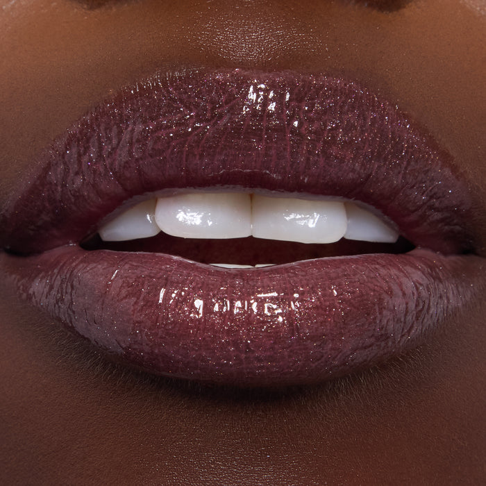 High-shine close-up of lips wearing ColourPop Ultra Glossy Lip in Ogress, a deep plum micro-shimmer gloss, reflecting light and showing a smooth, glassy finish on a model’s lips under studio lighting.