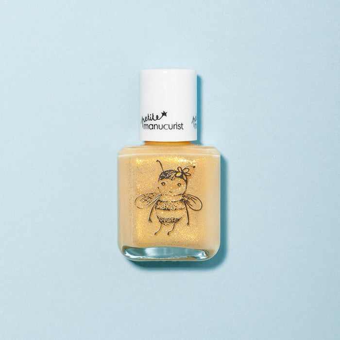 A bottle of Manucurist’s Pia the Bee kids' nail polish, featuring yellow glitter, a white cap, and a bee illustration on the front, sits on a light blue background. It’s made with naturally sourced ingredients.