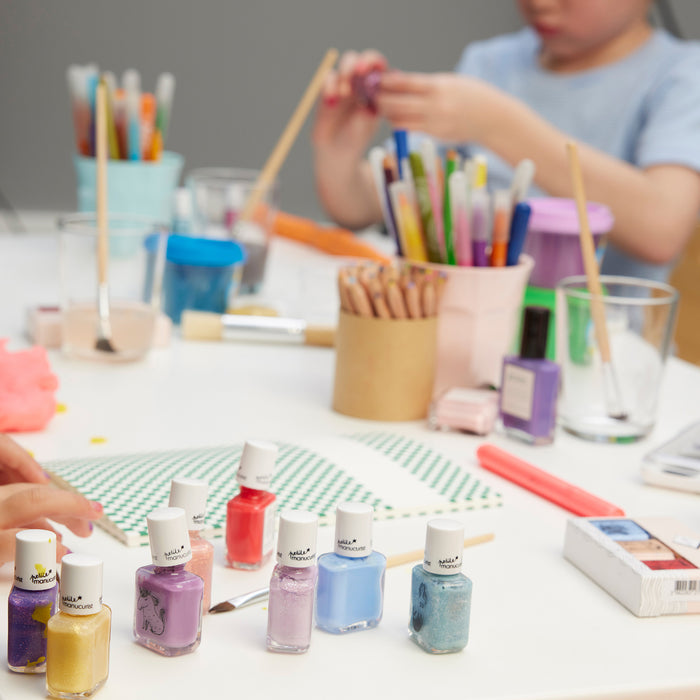 A table with Manucurist’s Pia the Bee kids' nail polish, colored pencils, paintbrushes, yellow glitter, and markers. In the background, a child works on a craft project.