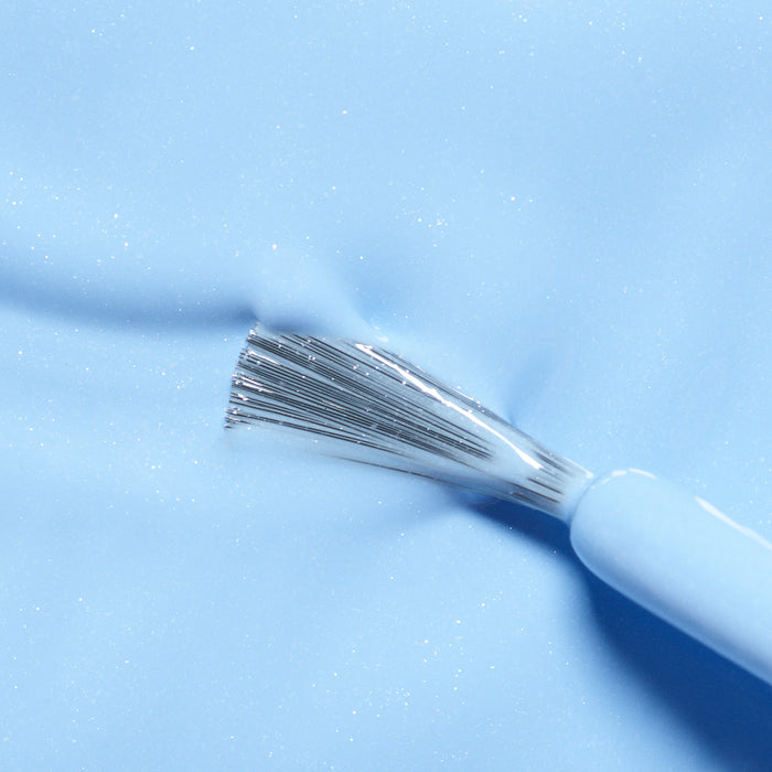 A close-up of a brush with silver bristles applying Manucurist’s Pomme the Fish, a light blue, non-toxic nail polish, on a smooth, glossy surface.