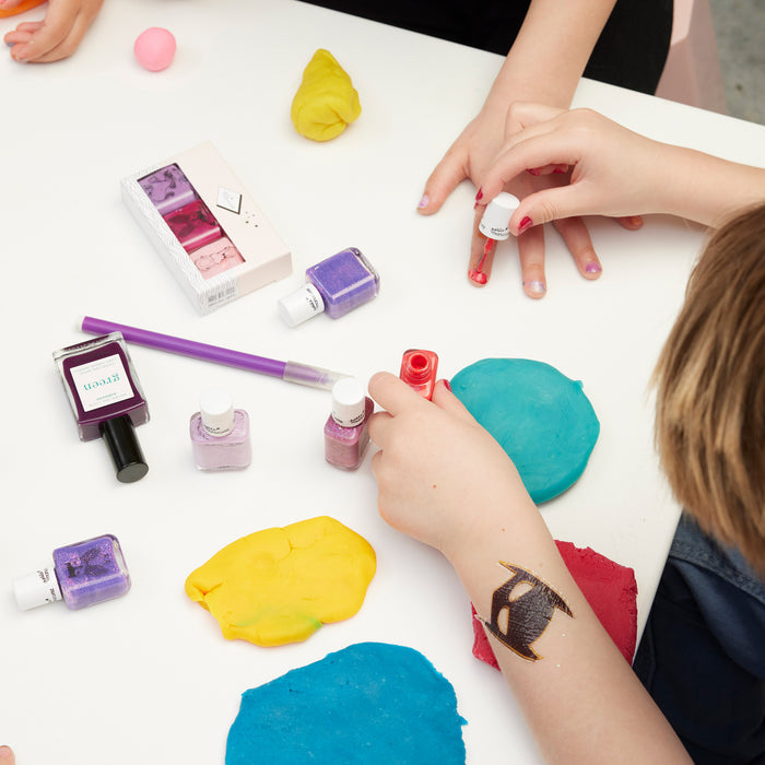 Children are painting their nails with Manucurist's Pomme the Fish, a colorful, non-toxic kids nail polish. Bottles of polish, brushes, and bright playdough are on the white table. One child sports a temporary tattoo on their arm.