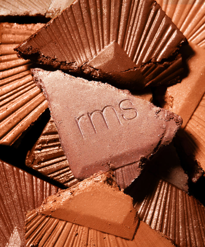 Close-up of various shades of broken pressed powder bronzer pieces, featuring one with "rms" embossed, highlighting the ReDimension Hydra Bronzer texture and natural ingredients.