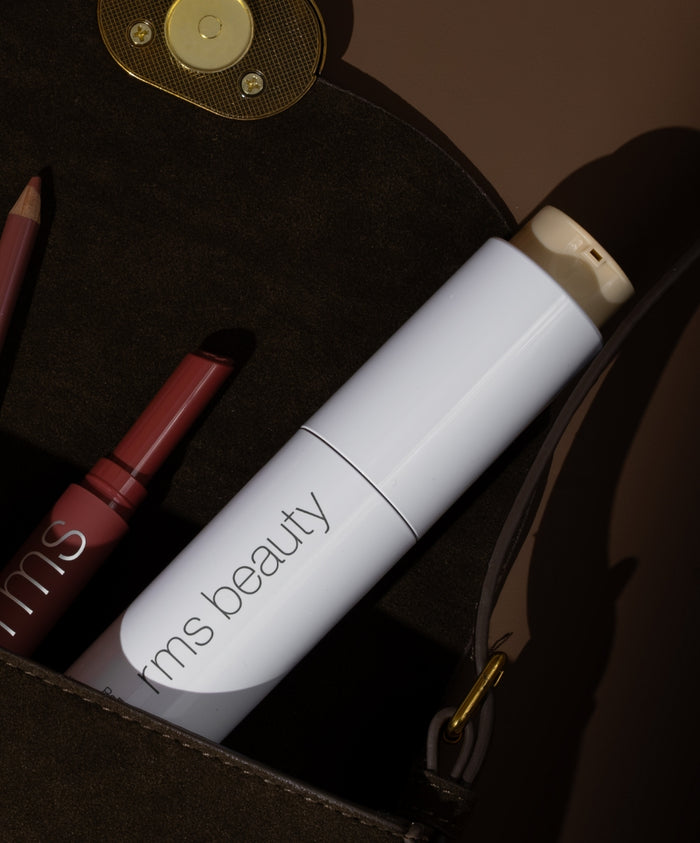 White sleek bottle of ReEvolve Natural Finish Liquid Foundation with rms beauty logo inside a brown bag alongside a makeup pencil and lipstick in warm lighting