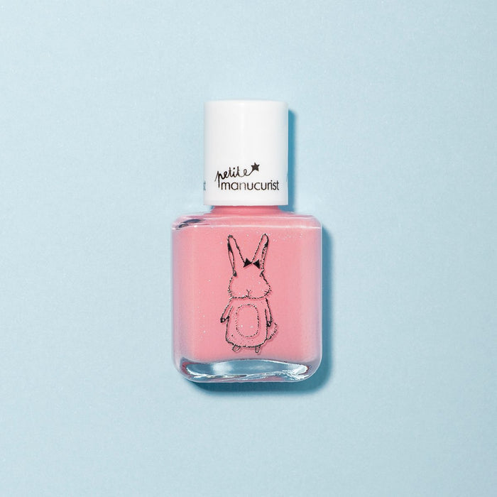 A bottle of Manucurist Rosie the Rabbit kids' nail polish in candy pink glitter, white cap, and a black bunny outline on the front sits on a light blue surface.