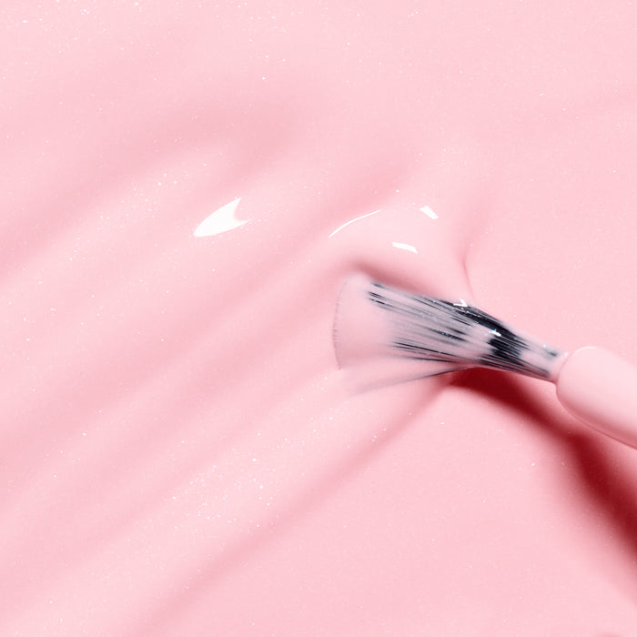 A close-up of a brush applying Rosie the Rabbit by Manucurist, a glossy candy pink glitter nail polish, creating soft, wavy lines on a smooth surface.