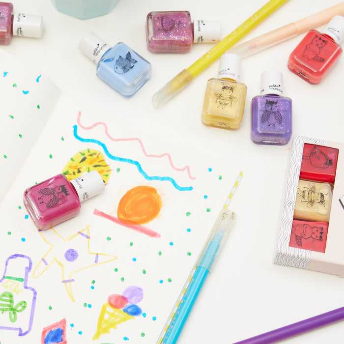 A sketchbook with colorful drawings is surrounded by Manucurist Scarlett the Owl kids' nail polish bottles featuring animal illustrations and naturally sourced ingredients, plus purple glitter pens and paintbrushes on a white surface.