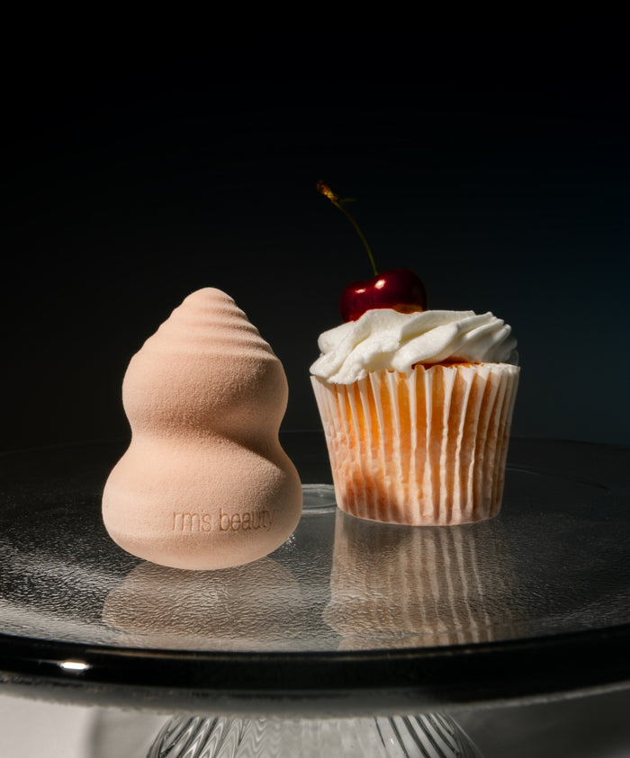 Skin2Skin Beauty Sponge next to a cherry-topped cupcake on a glass cake stand, highlighting the soft texture and ergonomic shape of the makeup sponge.