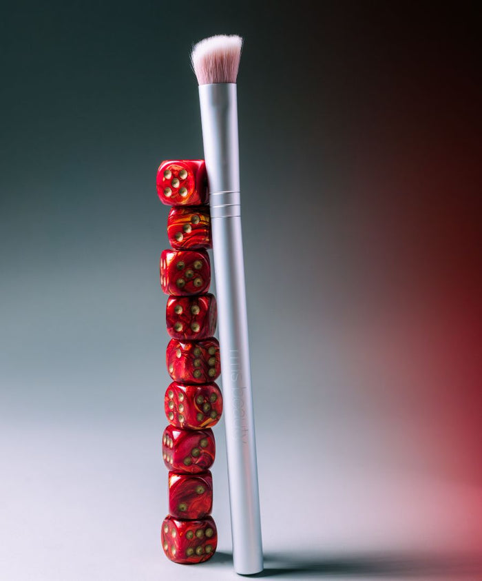 Skin2Skin Concealer Brush with soft pink bristles standing next to a vertical stack of red dice on a gradient gray and red background, highlighting clean beauty and cruelty free makeup.