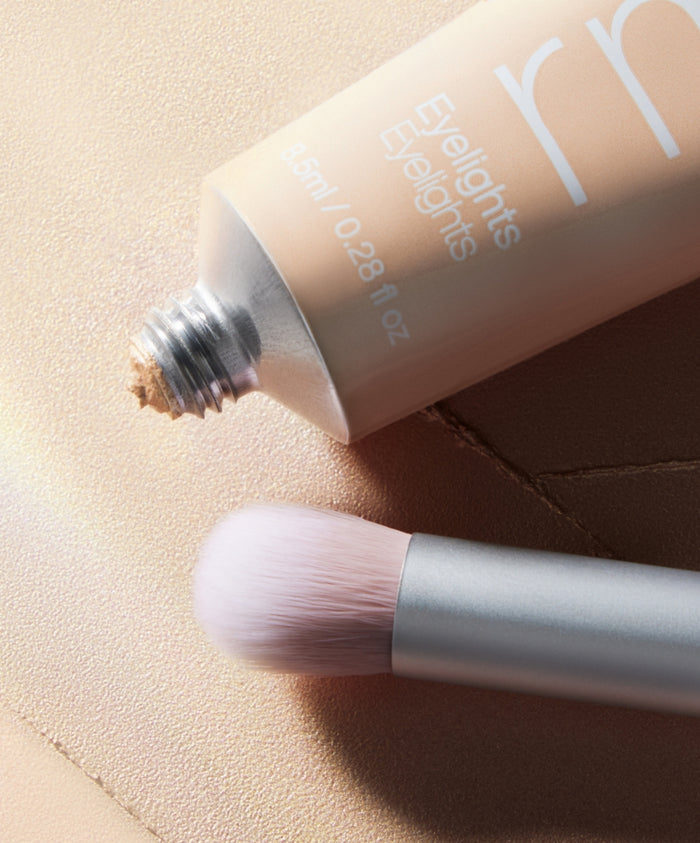Skin2Skin Everything Eye Polish Brush with soft bristles next to a tube of light beige cream makeup on a textured surface, highlighting clean beauty and natural ingredients.