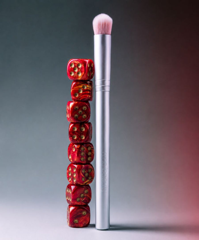 Skin2Skin Everything Eye Polish Brush with soft pink bristles and silver handle standing upright next to a stack of red dice, highlighting clean beauty and sustainable packaging.