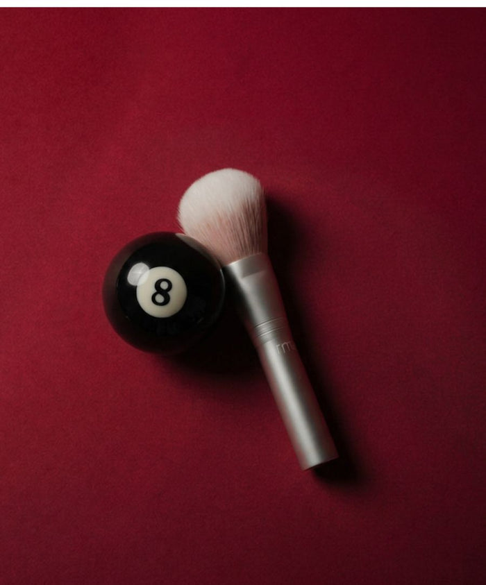 Skin2Skin Powder Blush Brush with soft white bristles and silver handle placed next to a black eight ball on a deep red background, highlighting clean beauty and cruelty free makeup.