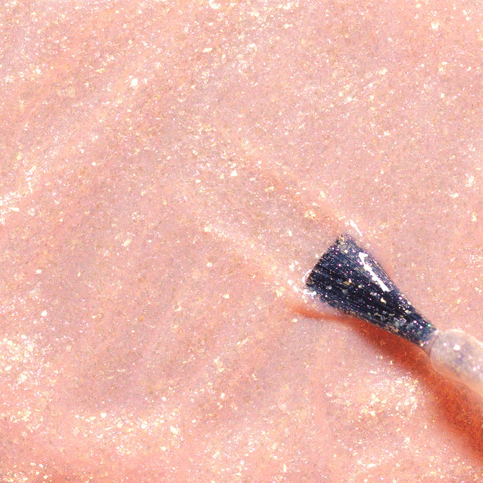 A close-up of a nail polish brush spreading Manucurist’s Stella the Starfish, a shimmery pale pink kids nail polish with gold glitter made from naturally sourced ingredients, over a smooth surface.