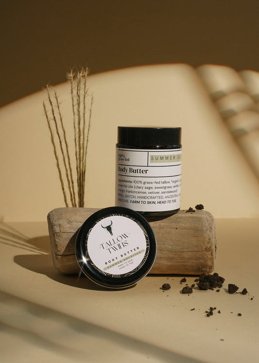 A jar of Summer Solstice Body Butter and its lid resting on driftwood with dried grasses.