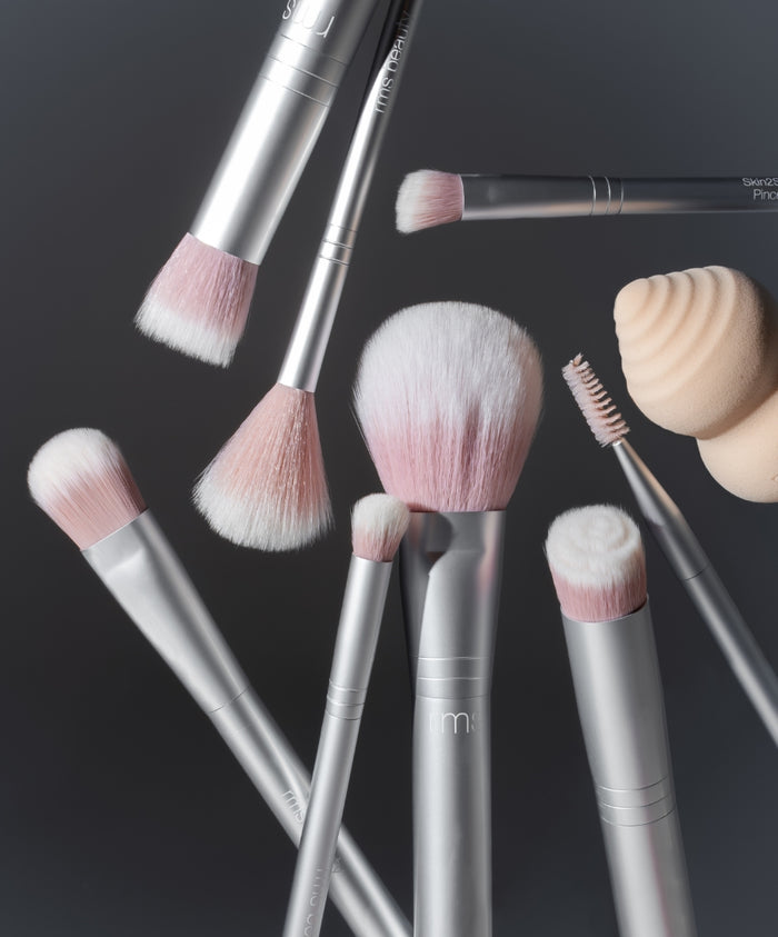 Close-up of various silver makeup brushes with soft pink and white bristles alongside a beige makeup sponge, arranged on a dark surface representing The Artist Toolkit.